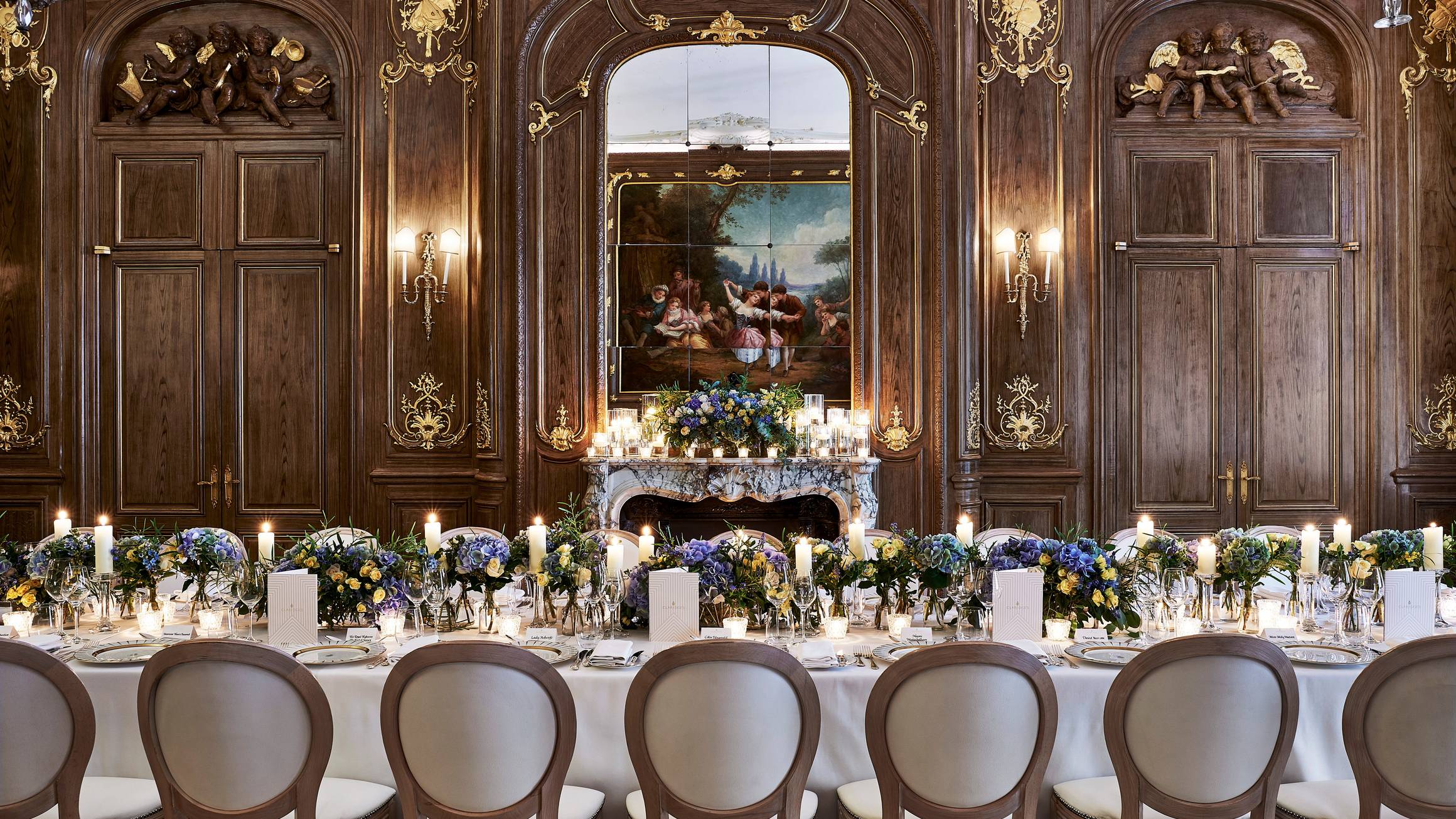 A lavishly set dining table in an opulent wood-paneled room adorned with gold details, featuring elegant floral arrangements, glowing candles, and a grand marble fireplace beneath a classical painting.