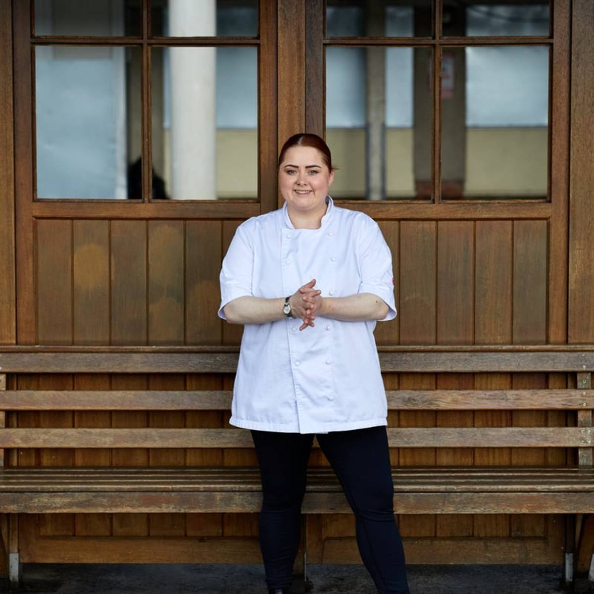 Chef in a white jacket and black boots stands confidently in front of a rustic wooden backdrop, smiling warmly.