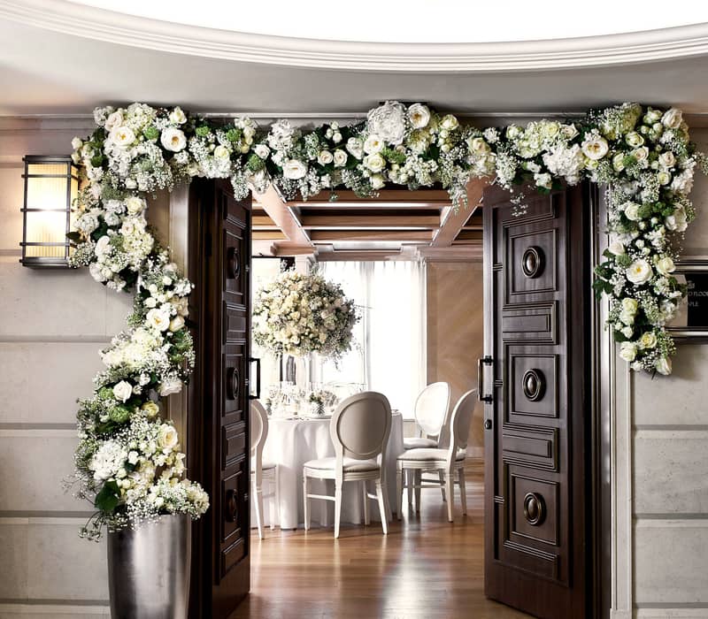 An elegant entryway adorned with a lavish floral garland of white roses and greenery, leading into a beautifully decorated dining space with round tables, white chairs, and stunning floral centerpieces under warm, natural light.