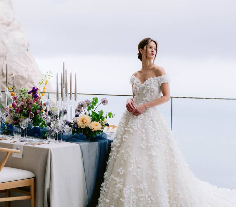 A bride in an intricate floral gown poses gracefully outdoors at Maybourne Riviera, surrounded by modern architecture.