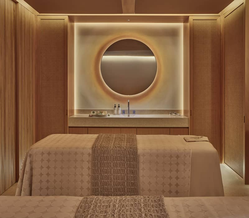 Elegant spa treatment room with two massage tables, warm wood paneling, soft lighting, and a circular backlit mirror above a minimalist counter with skincare products.