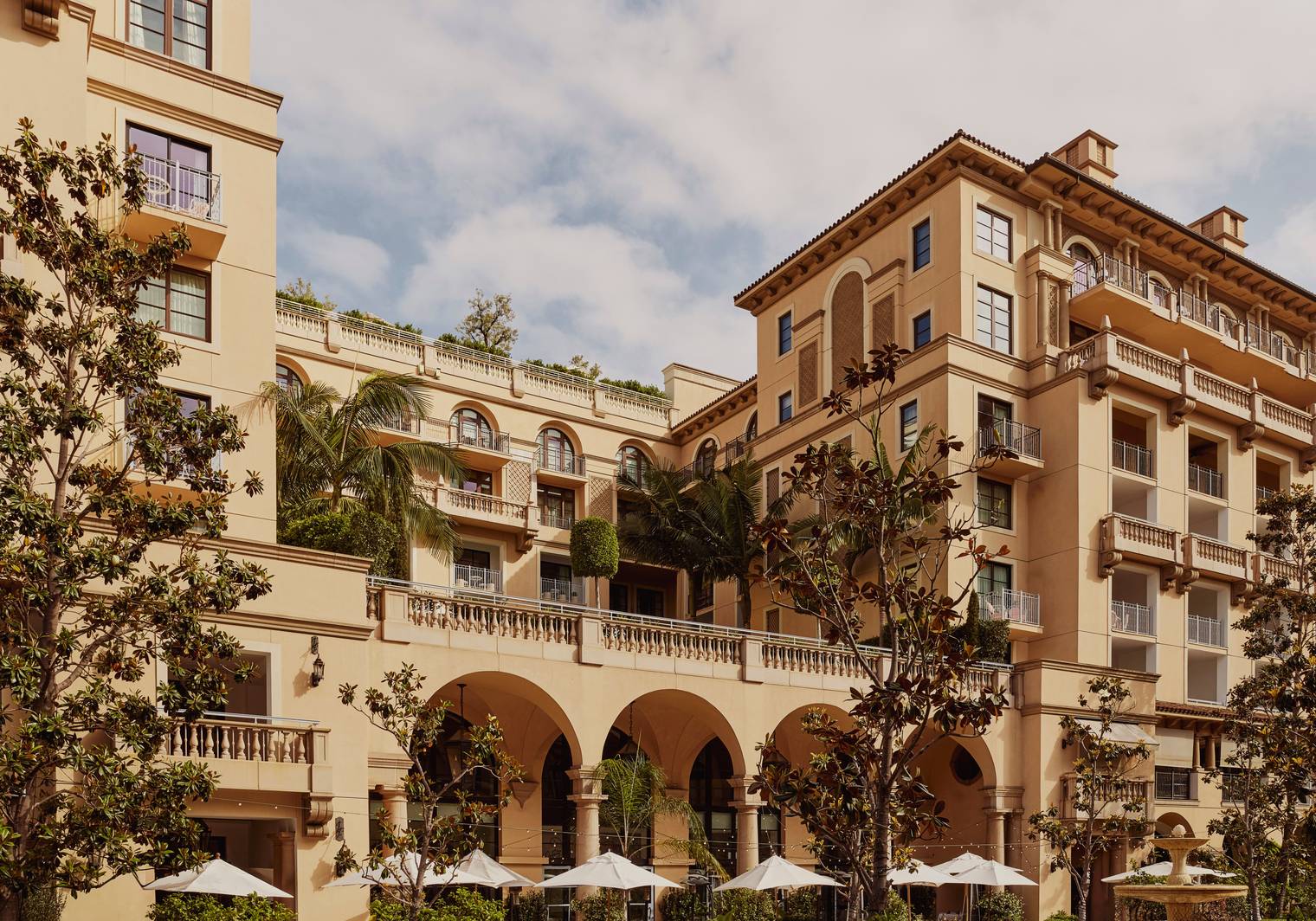 A luxurious Mediterranean-style hotel with arched balconies, lush greenery, and an outdoor terrace adorned with umbrellas and palm trees.