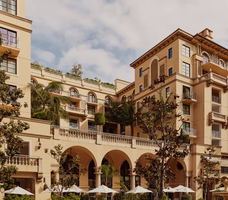 A luxurious Mediterranean-style hotel with arched balconies, lush greenery, and an outdoor terrace adorned with umbrellas and palm trees.