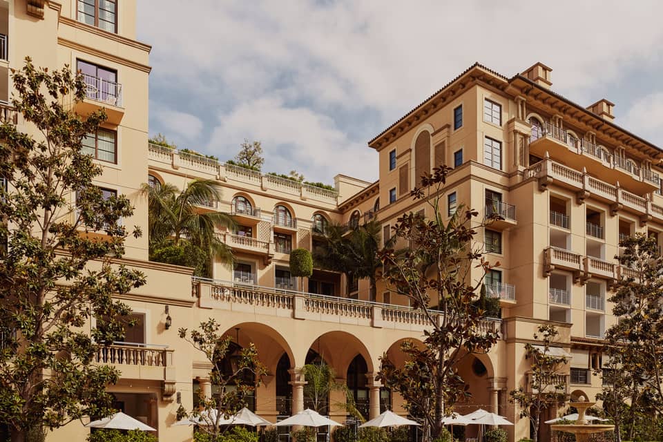 A luxurious Mediterranean-style hotel with arched balconies, lush greenery, and an outdoor terrace adorned with umbrellas and palm trees.