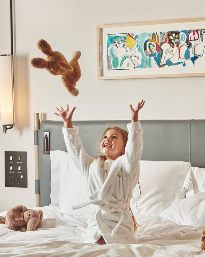 A joyful child in a white robe tossing a plush toy on a luxurious bed, with playful art adorning the wall.