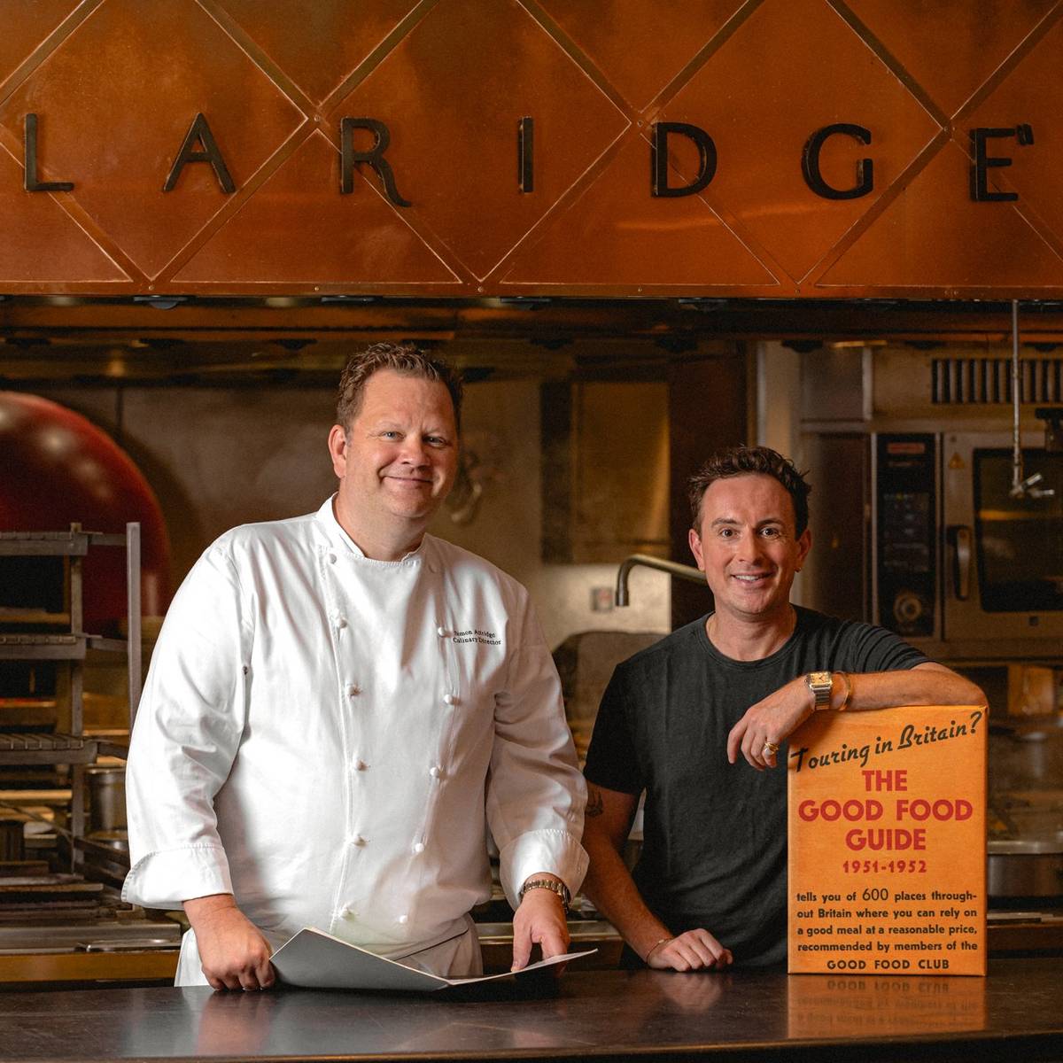 Two chefs standing in Claridge’s kitchen, one holding a vintage Good Food Guide, with copper decor and pizza oven behind.