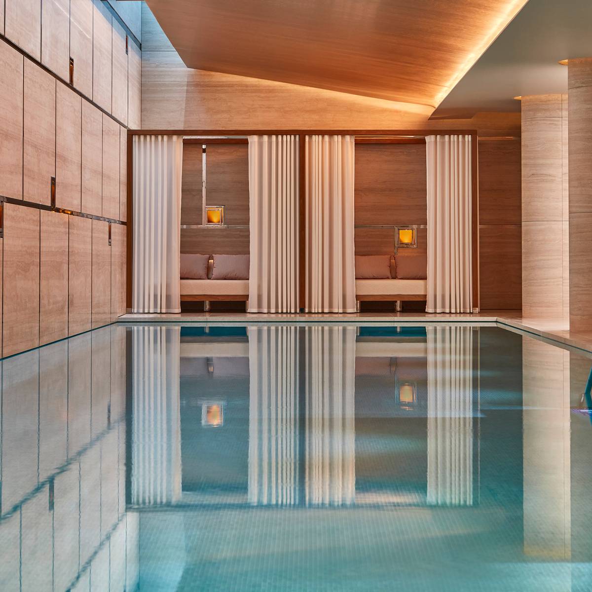 Tranquil indoor pool with stone walls, soft lighting, and curtained poolside cabanas creating a calming retreat