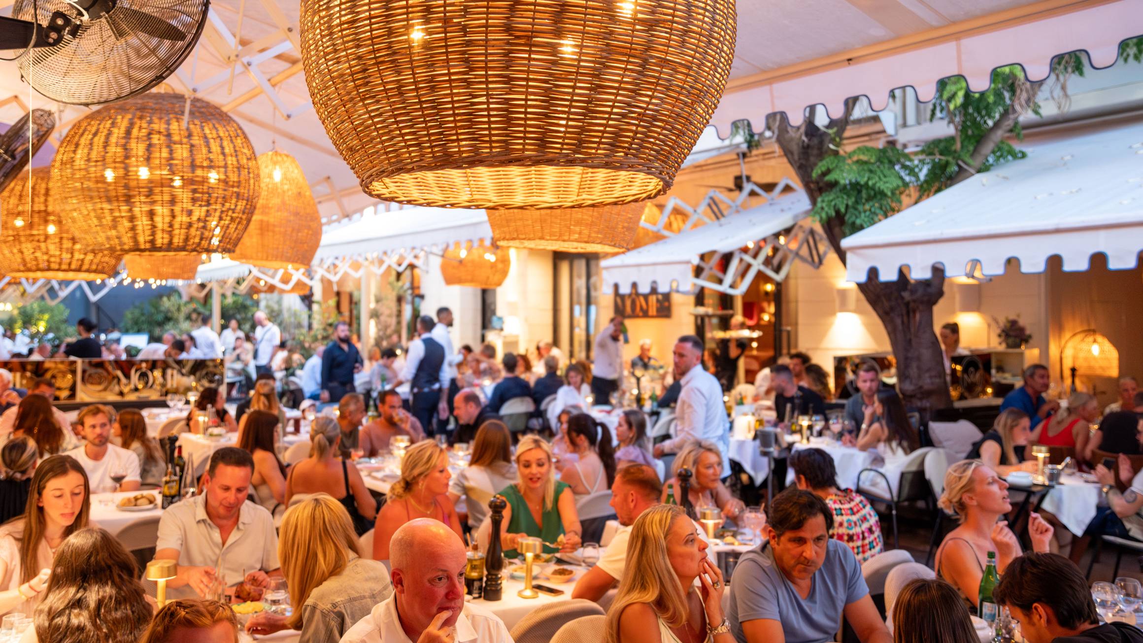An elegant, bustling outdoor restaurant with wicker pendant lights, lively conversations, and diners enjoying a sophisticated ambiance under a canopy, evoking a warm Mediterranean charm.