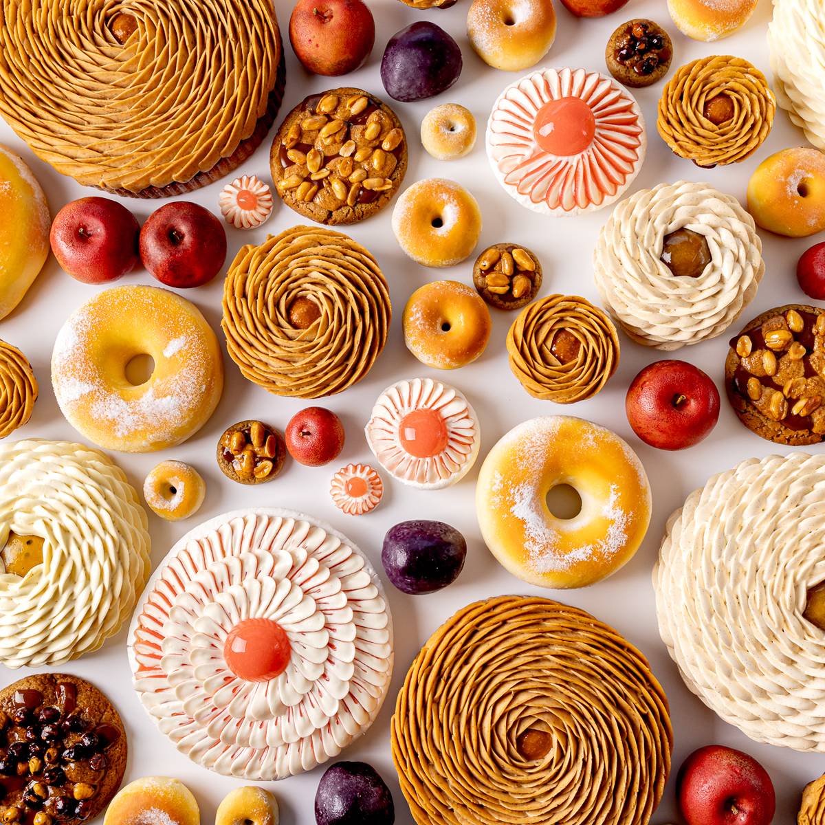 Assortment of intricately crafted pastries and fruit-shaped desserts in vibrant colours, arranged on a white background in a visually striking, artistic flat lay.