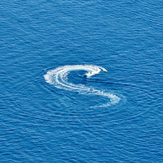 A speedboat creates a striking "S" pattern of white wake across the vibrant, endless expanse of deep blue ocean.