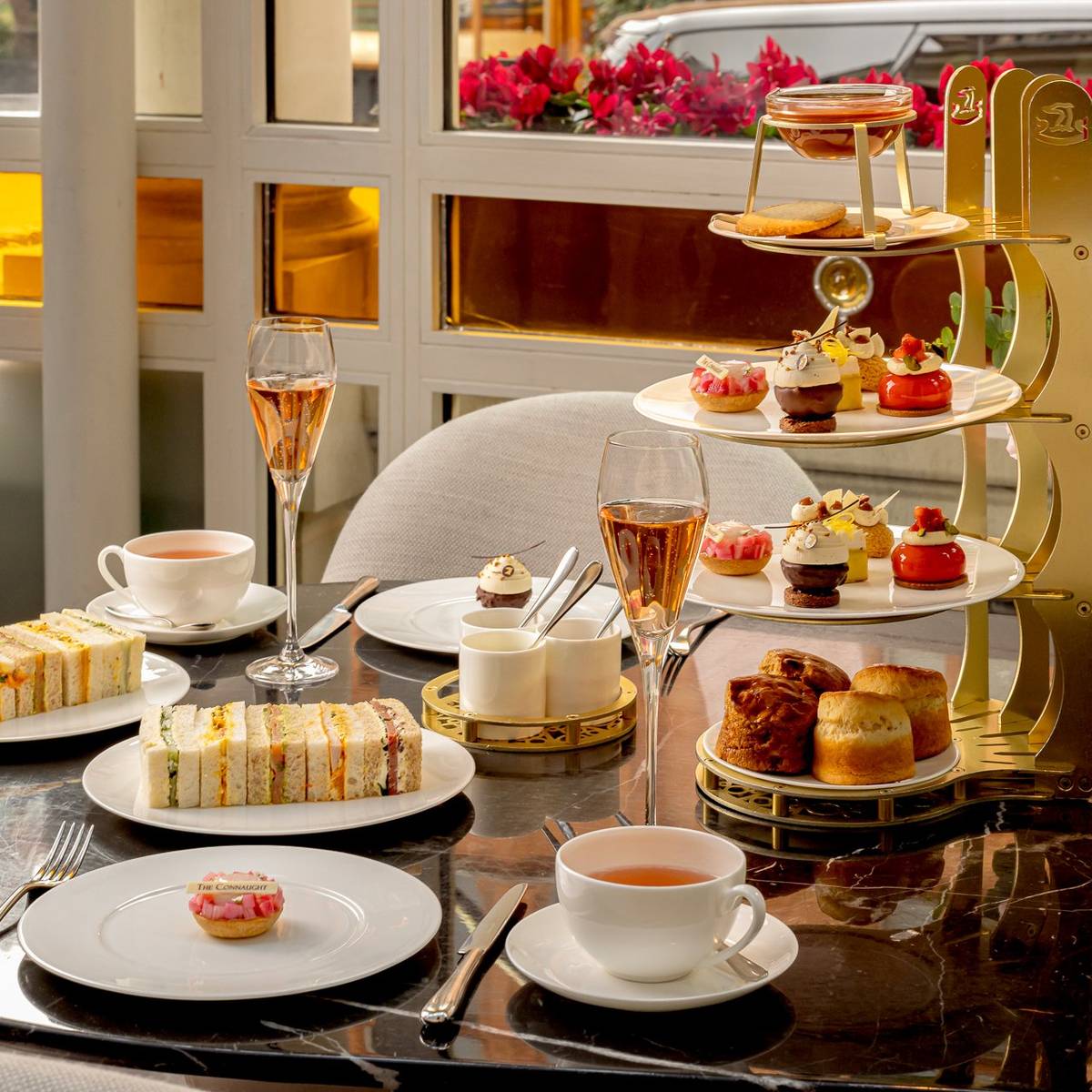 Afternoon tea set on a marble table with finger sandwiches, pastries, scones, tea, rosé champagne, and a vase of fresh flowers.