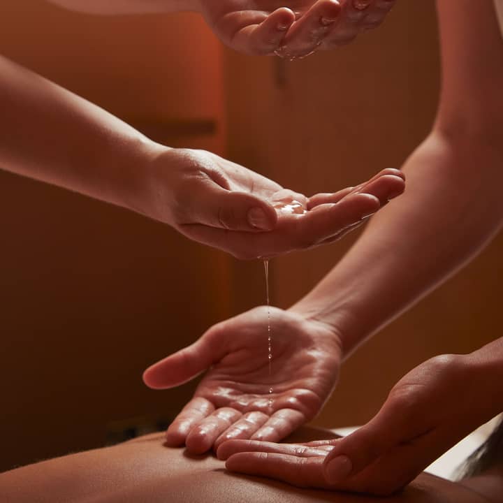 Hands gently pouring warm oil during a spa massage treatment in a softly lit, serene setting.