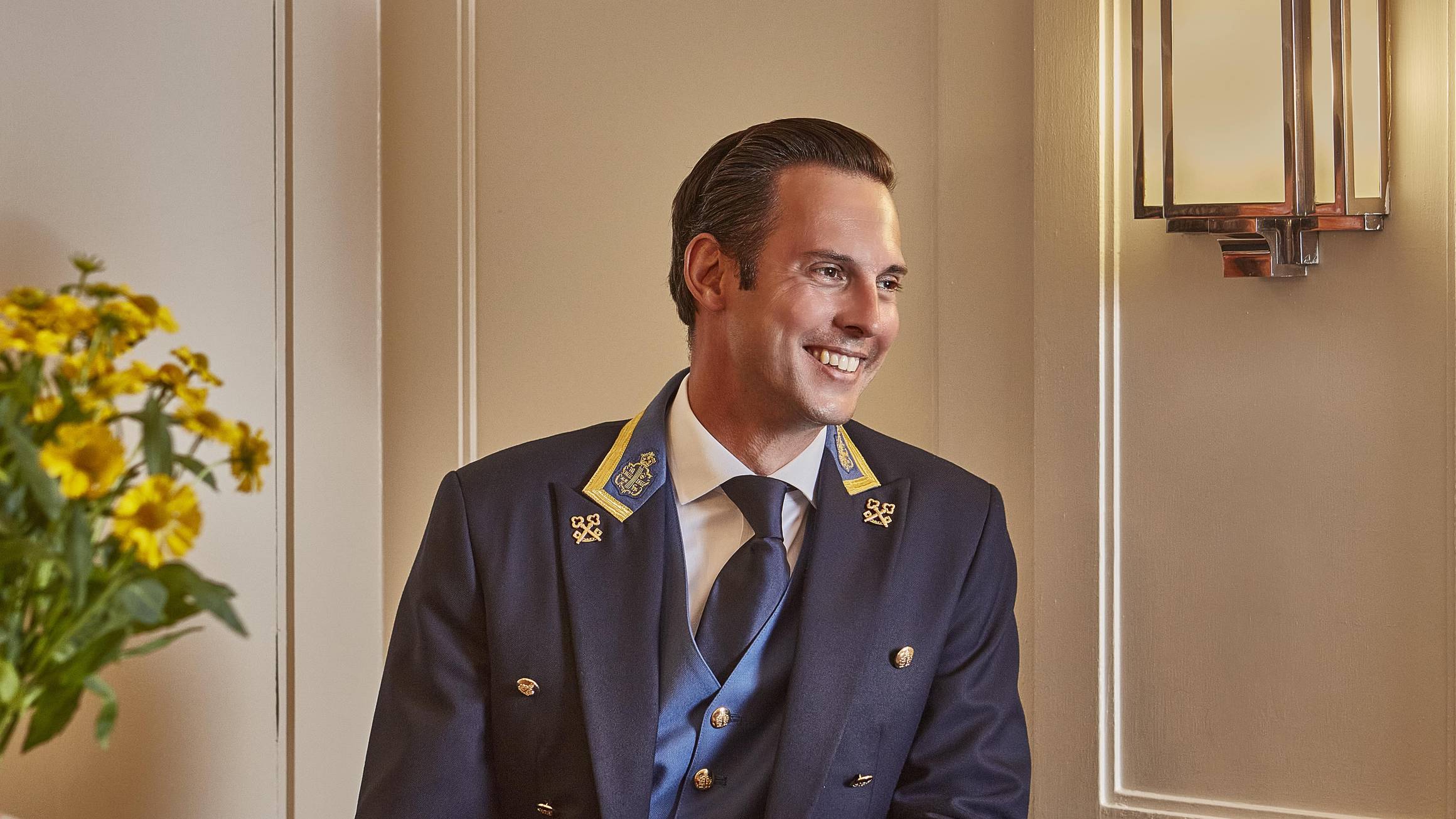 A smiling concierge in an elegant uniform, seated at a polished desk with warm lighting and fresh yellow flowers.