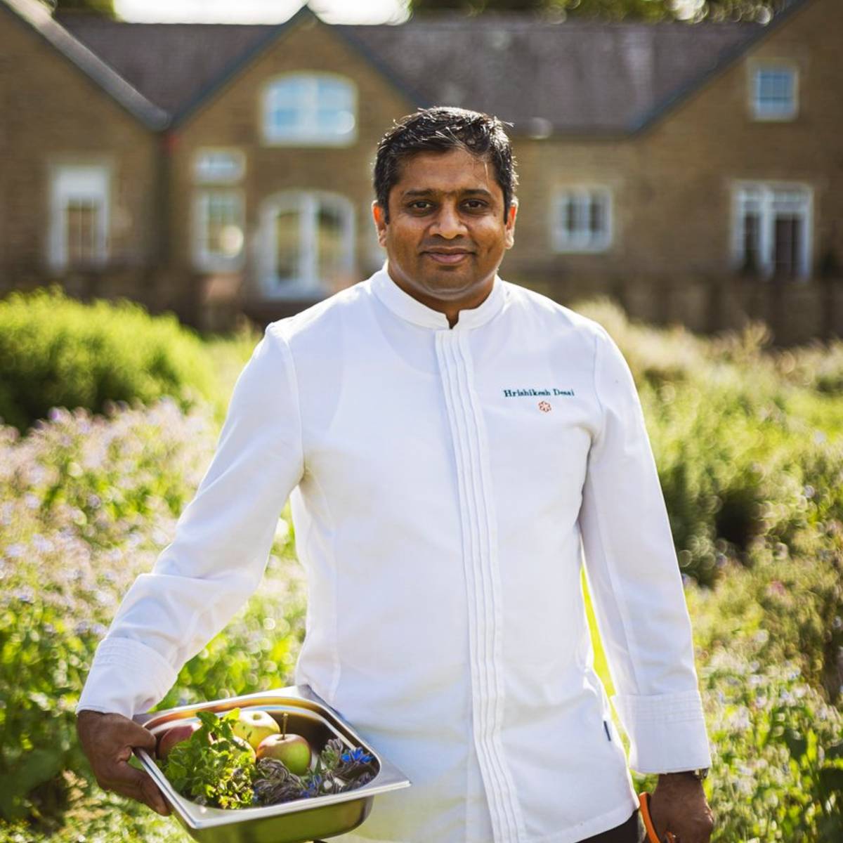 Chef in a crisp white jacket holding a tray of fresh produce, standing in a lush garden with a manor house.