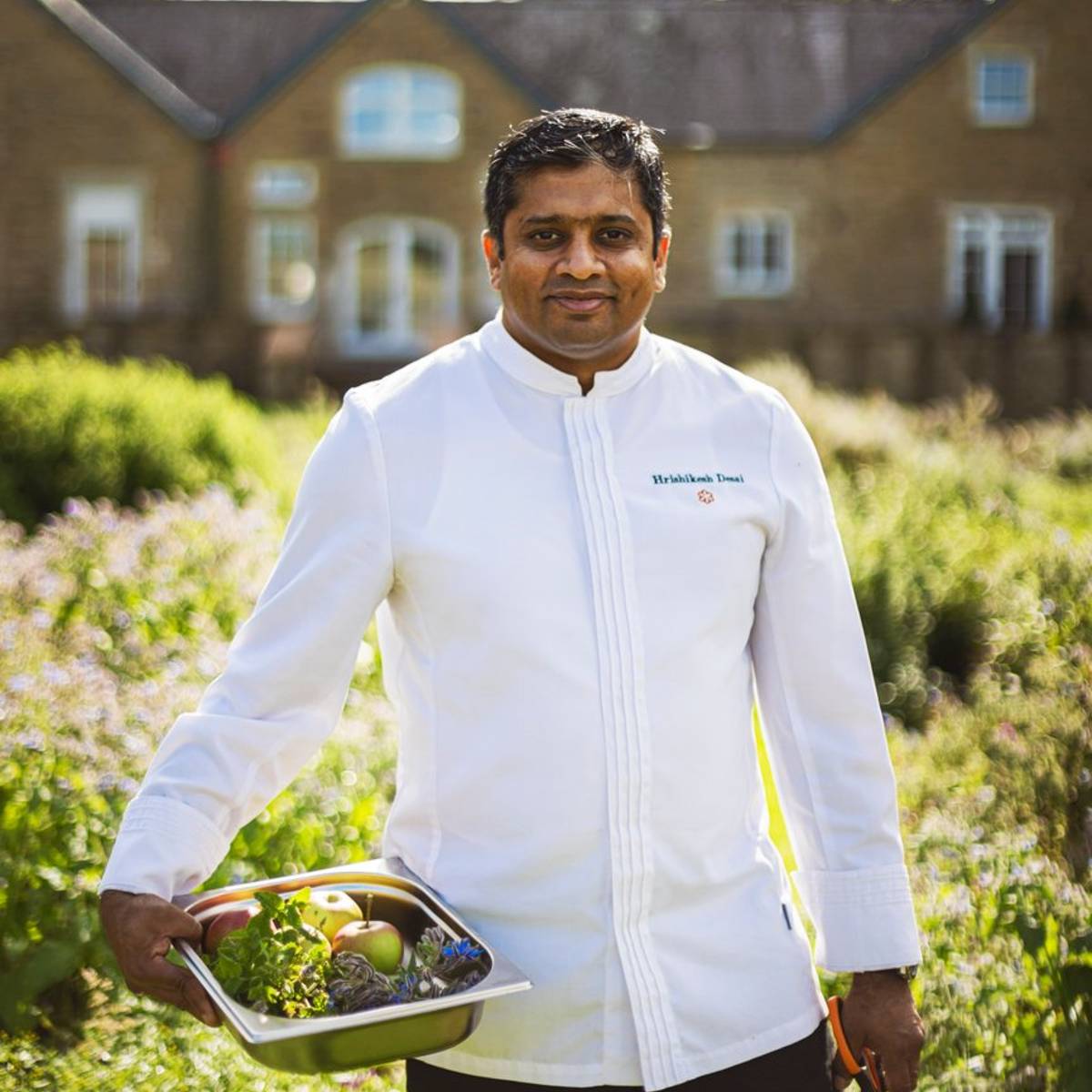 Chef in a crisp white jacket holding a tray of fresh produce, standing in a lush garden with a manor house.