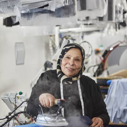 Smiling seamstress wearing a patterned headscarf steams fabric in a garment workshop, surrounded by clothing and equipment. Measuring tape hangs around her neck.