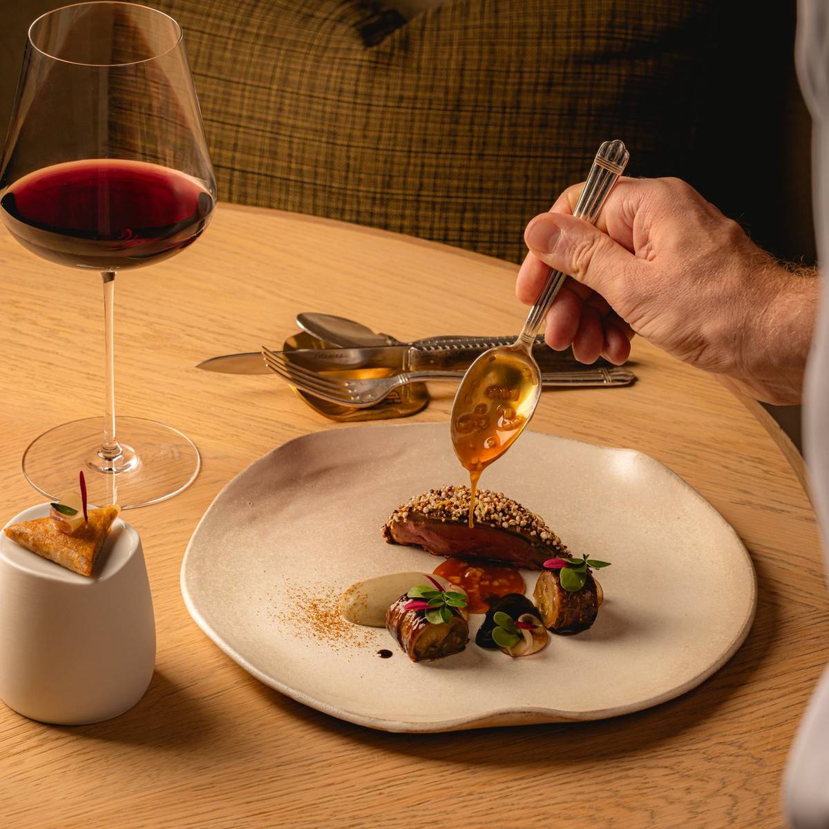 A Chef hand is carefully plating a gourmet meat dish, pouring sauce over vibrant ingredients on a round white dish, sitting on a round wooden table. A glass of red wine and cutlery are also on the table. A brown cushion and chair sit in the background.