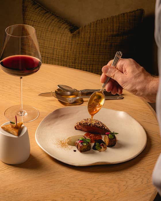 A Chef hand is carefully plating a gourmet meat dish, pouring sauce over vibrant ingredients on a round white dish, sitting on a round wooden table. A glass of red wine and cutlery are also on the table. A brown cushion and chair sit in the background.