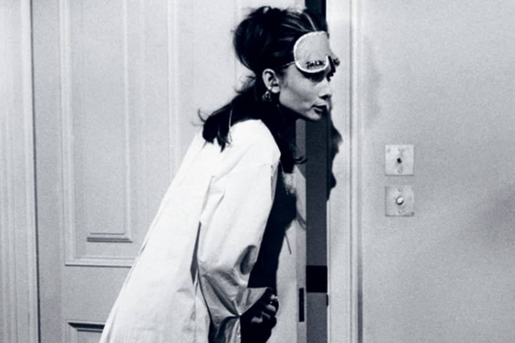 A woman wearing a sleep mask and oversized shirt leans curiously through a partially opened door in a dimly lit room.