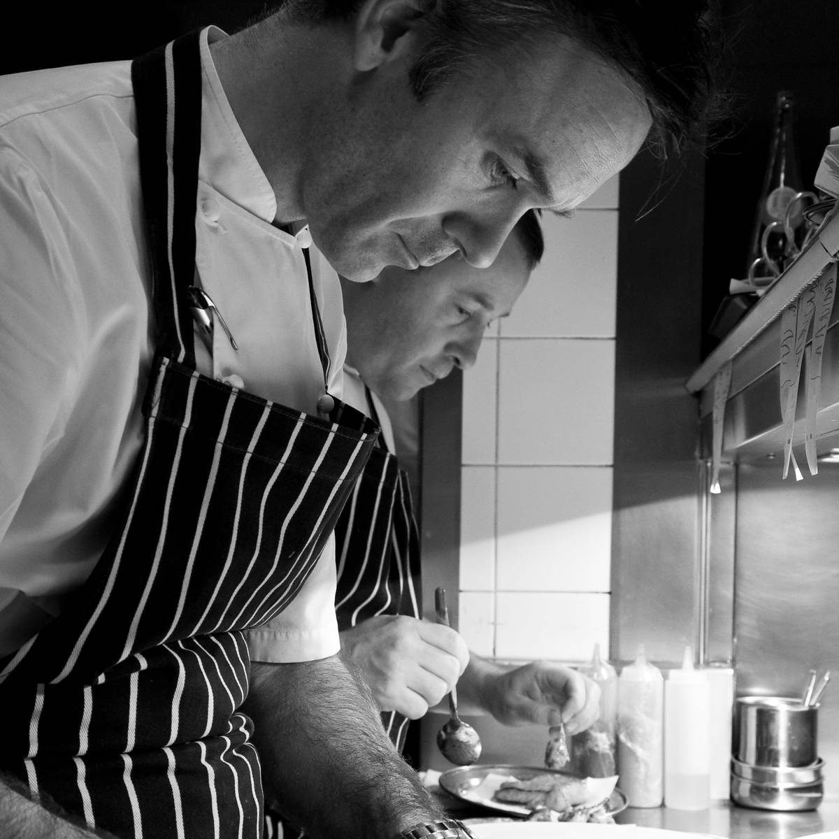 A black-and-white image of two chefs in striped aprons meticulously plating dishes in a professional kitchen.
