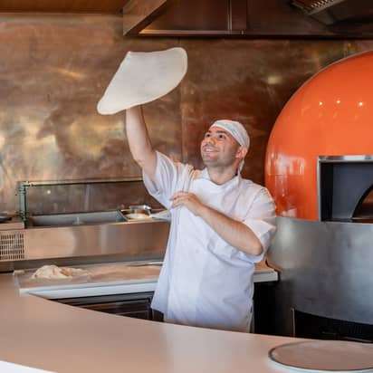 A cheerful chef tossing pizza dough in an artisan kitchen with a copper interior and a blazing orange oven.