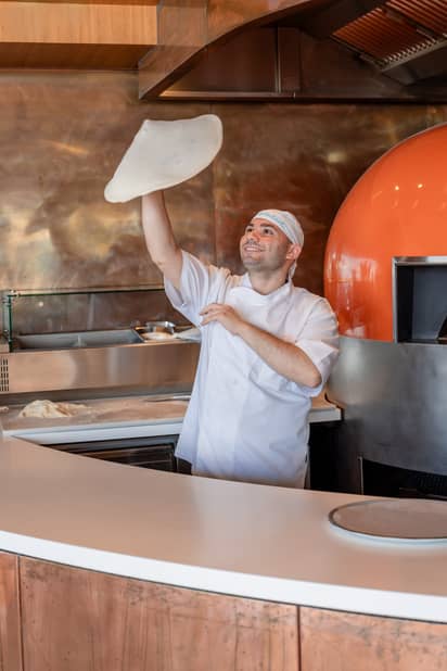 A cheerful chef tossing pizza dough in an artisan kitchen with a copper interior and a blazing orange oven.