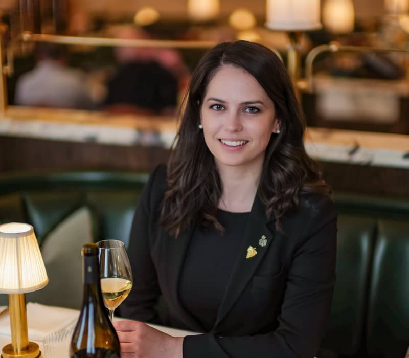 A smiling woman in a black blazer sits at an elegant restaurant table with wine, a glass, and soft lighting.