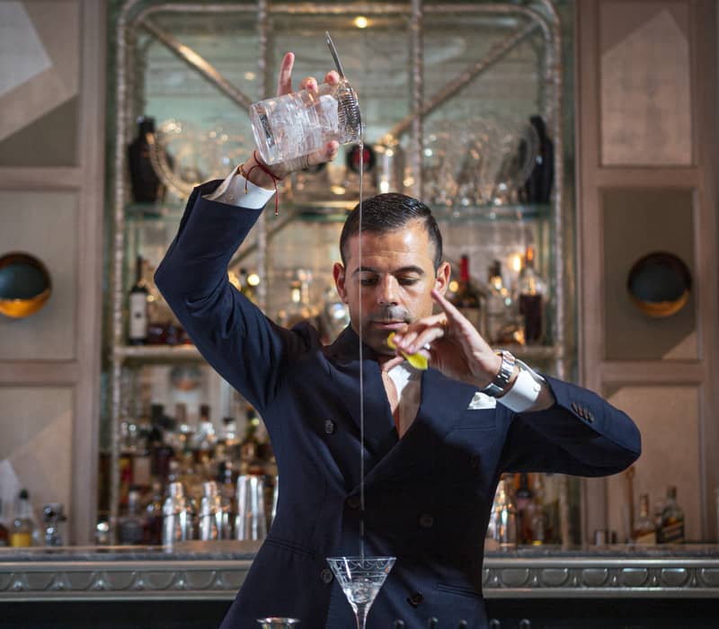 Bartender in a tailored suit elegantly pouring a cocktail from height, showcasing precision and skill in a luxurious bar setting.