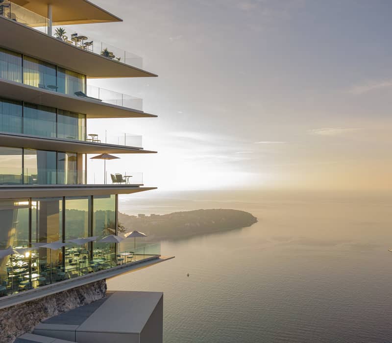 Luxury seafront residence with tiered balconies, glass walls, and sunset reflections over the Mediterranean coastline.