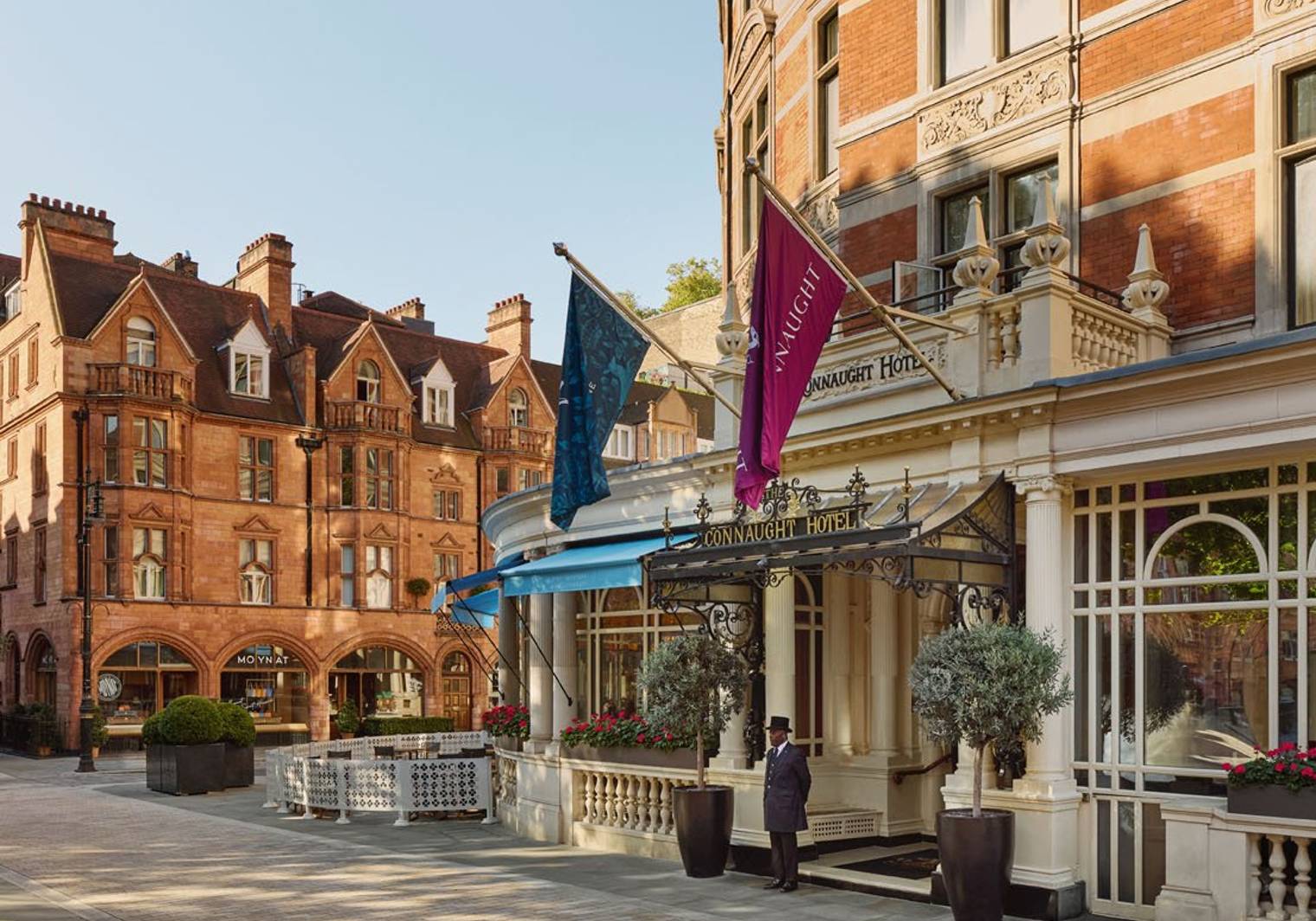The elegant exterior of The Connaught Hotel, featuring red-brick architecture, flags, and a doorman under a decorative entrance.