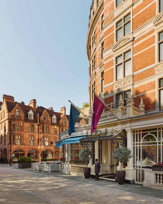 The elegant exterior of The Connaught Hotel, featuring red-brick architecture, flags, and a doorman under a decorative entrance.