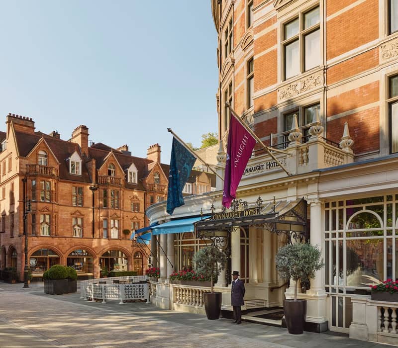The elegant exterior of The Connaught Hotel, featuring red-brick architecture, flags, and a doorman under a decorative entrance.