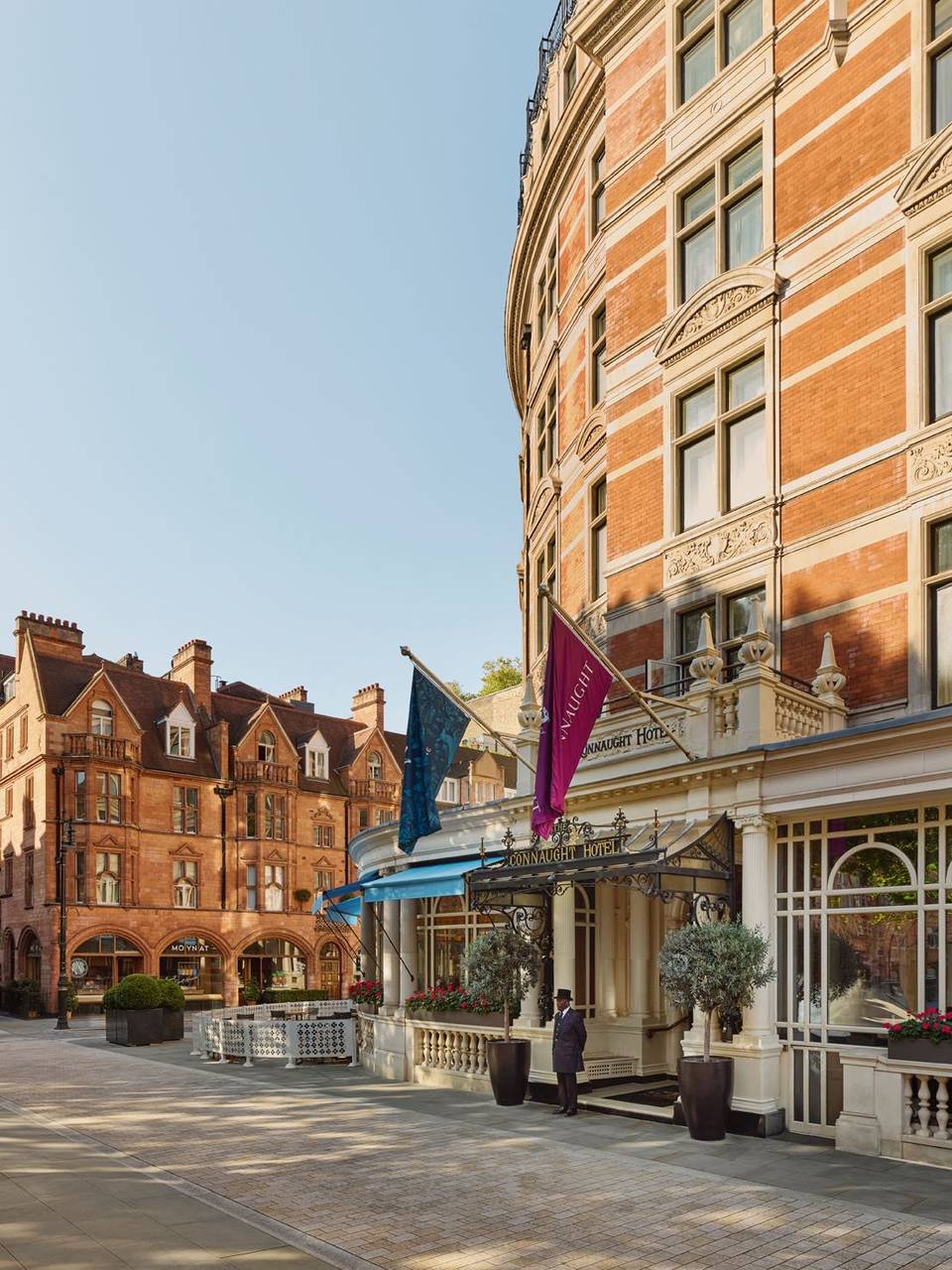 The elegant exterior of The Connaught Hotel, featuring red-brick architecture, flags, and a doorman under a decorative entrance.