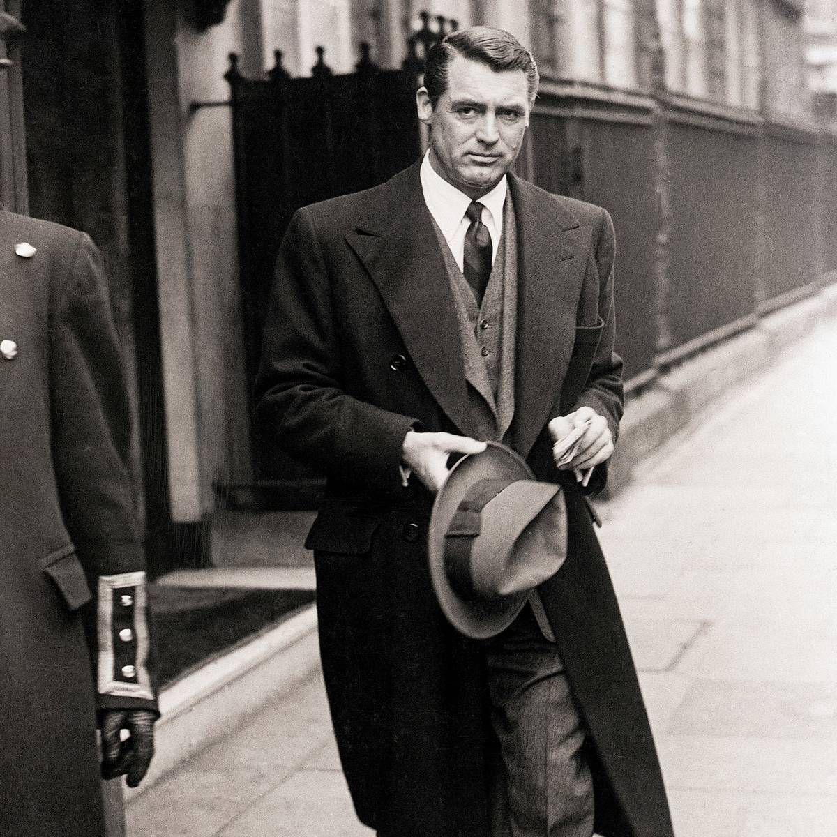 Cary Grant in overcoat and suit walking on city sidewalk, holding a fedora, with a uniformed doorman partially visible.