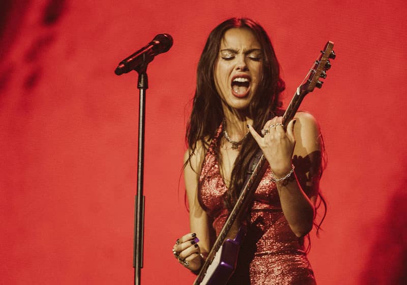 Olivia Rodrigo performs live, singing passionately with a purple guitar against a bold red backdrop, wearing a shimmering red outfit