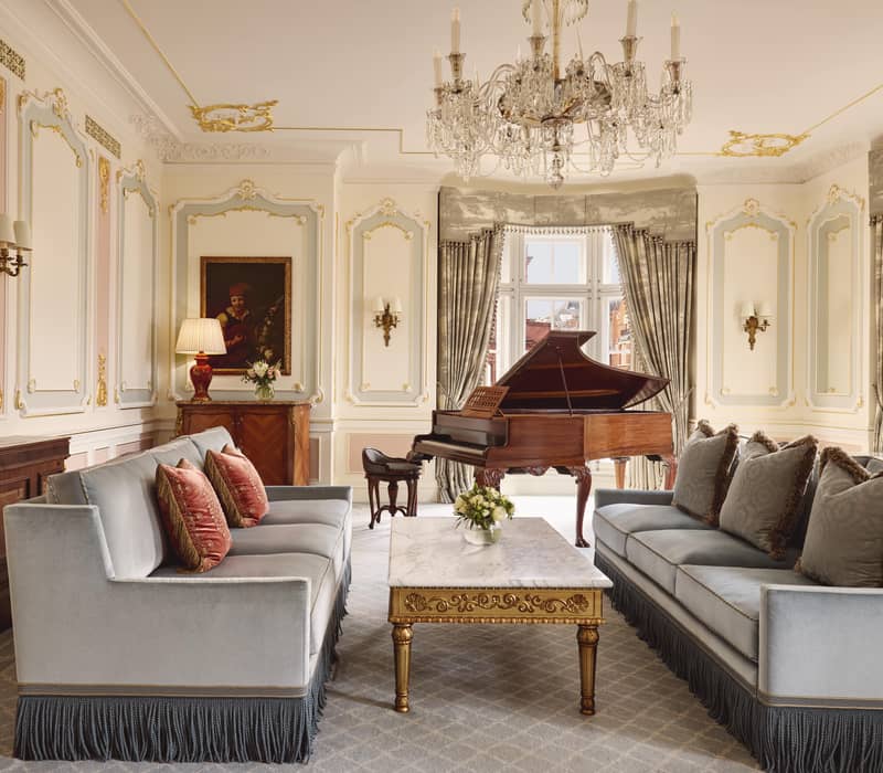 Opulent lounge in Claridge’s Prince Alexander Suite with grand piano, crystal chandelier, ornate gold detailing, and plush sofas with fringed skirts.