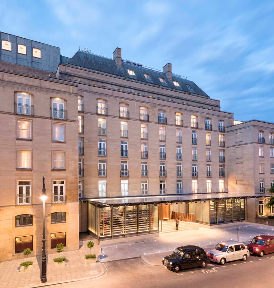 an iconic luxury hotel entrance with warm lighting, sleek glass canopy, and classic London taxis.