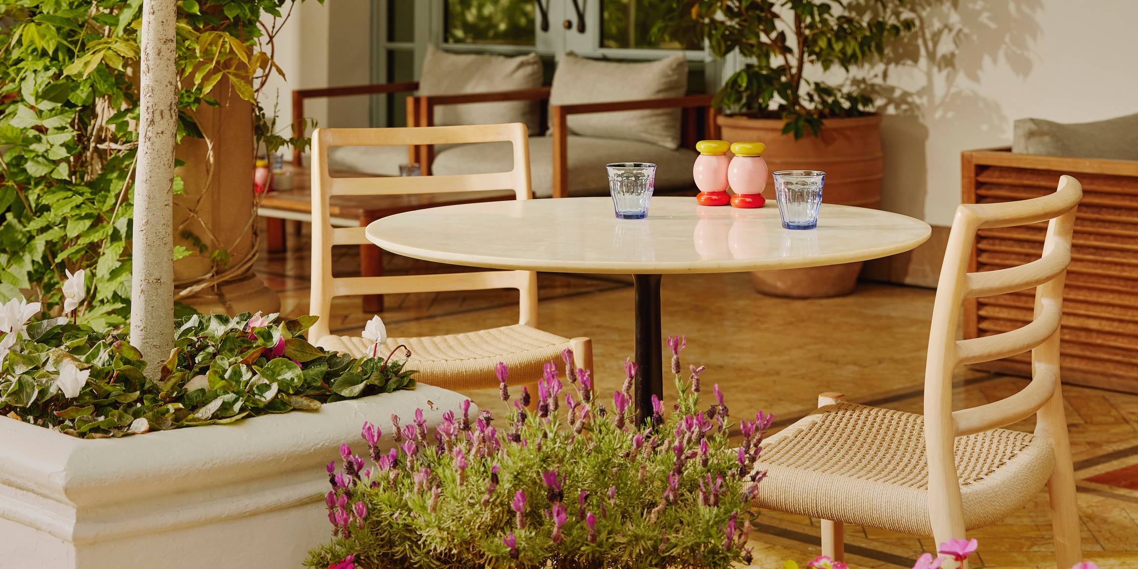 A serene outdoor café setting featuring a round table, woven chairs, vibrant potted flowers, and lush greenery under warm sunlight.