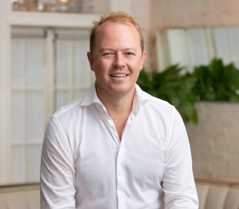 Smiling man in a white shirt and navy trousers seated in a light, modern restaurant interior.