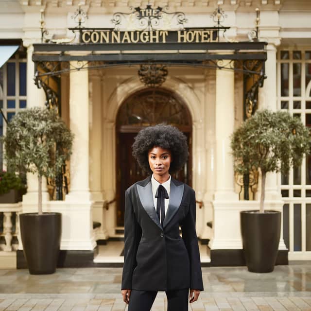 A confident individual in a tailored black suit with satin lapels stands gracefully outside The Connaught Hotel's grand entrance,