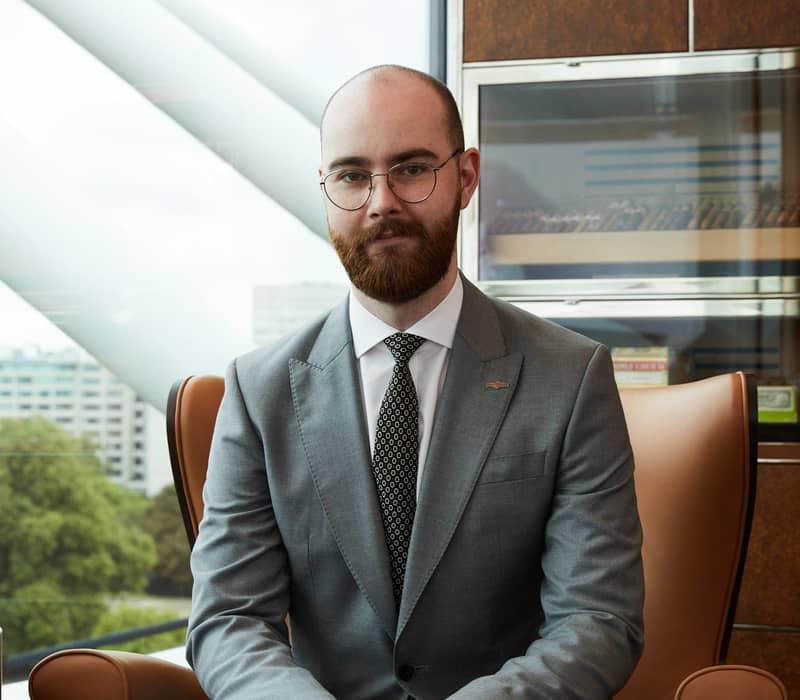 Man in a grey suit seated in a modern lounge with city views, looking directly at the camera.