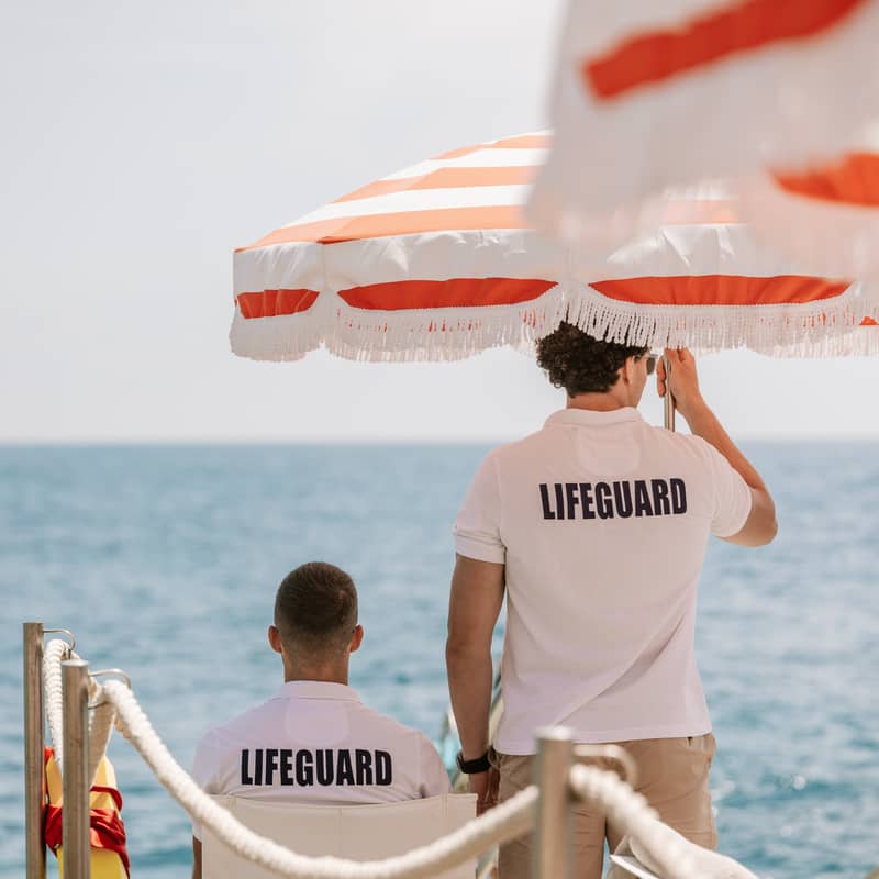 Lifeguards under orange-striped umbrellas overlooking a serene blue sea, ensuring safety on a sunny coastal day.