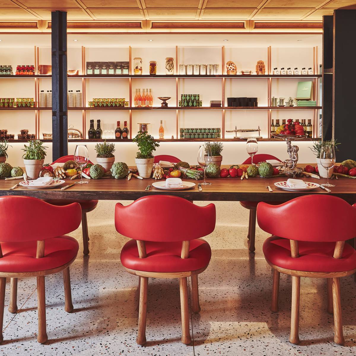 Long wooden dining table with red chairs, fresh produce, and herbs, set against open pantry shelves in warm lighting.