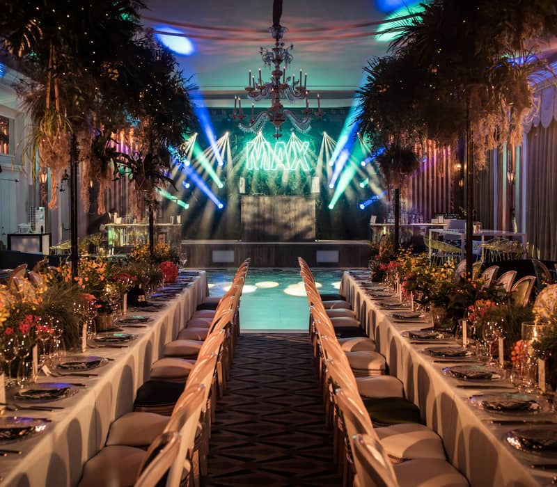 Lavish event space with long banquet tables, lush foliage, and vibrant stage lighting for a glamorous celebration.
