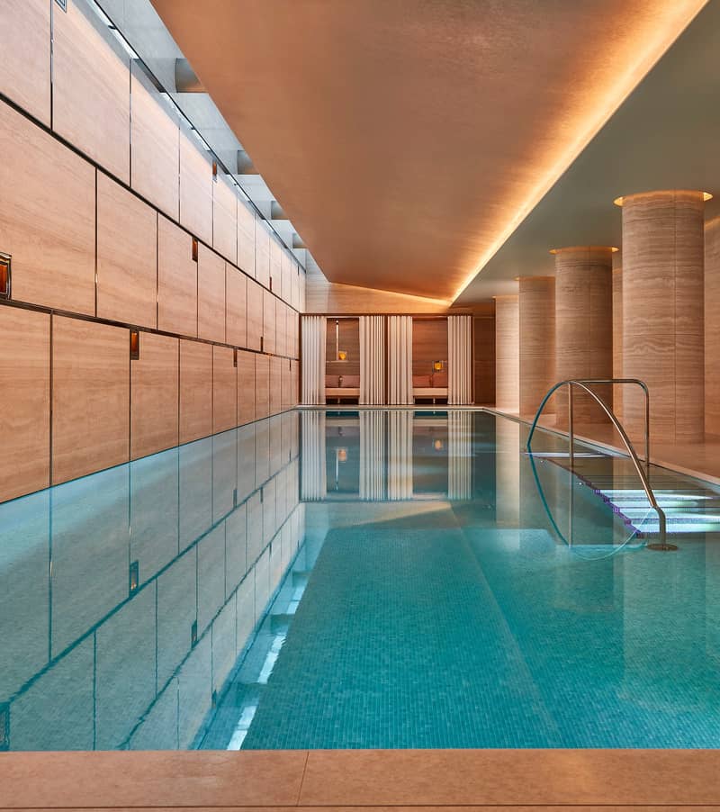 A serene indoor swimming pool with warm-toned stone walls, soft lighting, and elegant columned architecture creating a tranquil retreat.
