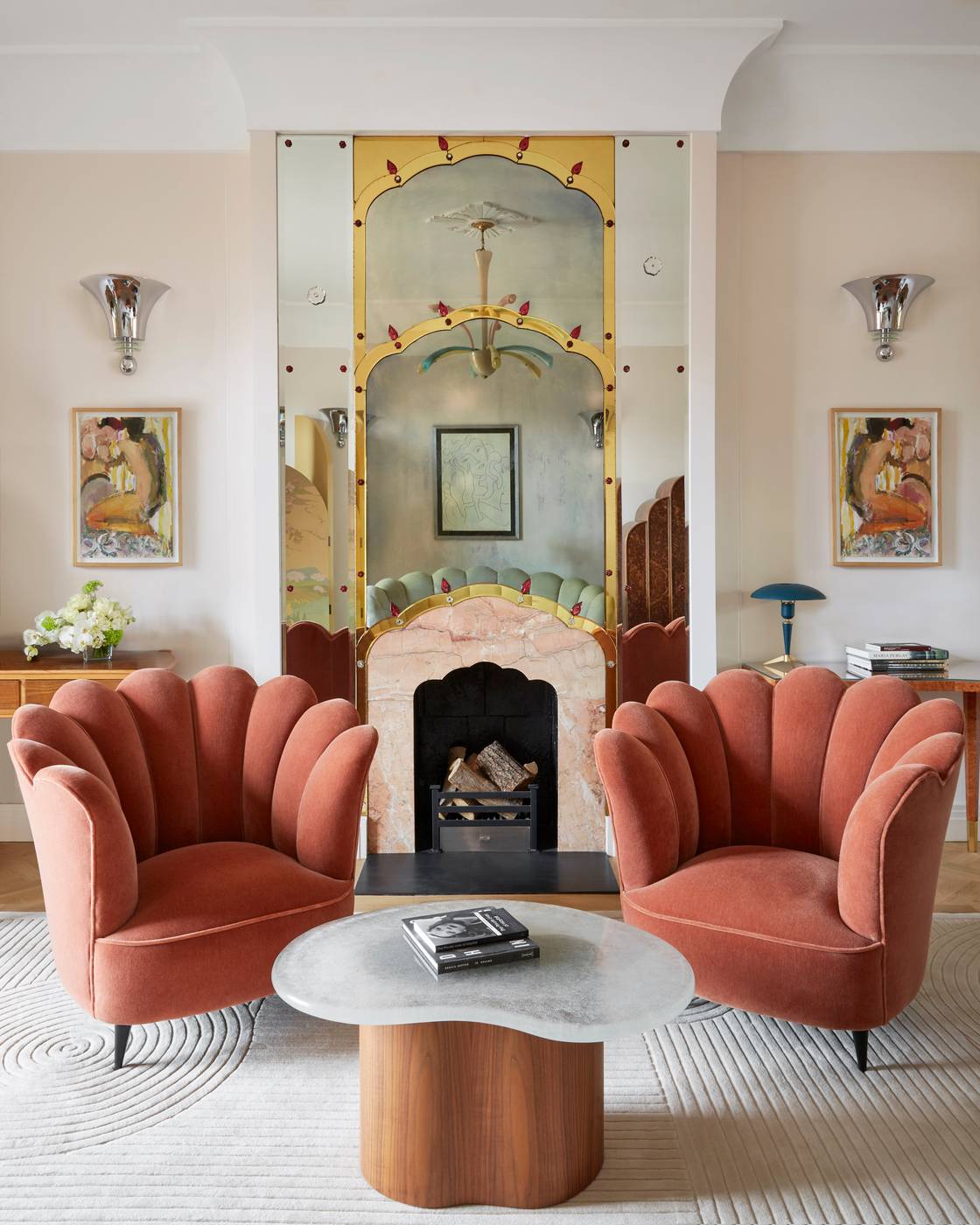 Two scalloped velvet armchairs and a sculptural coffee table face a decorative fireplace with mirrored Art Deco surround in an elegant, light-filled suite.