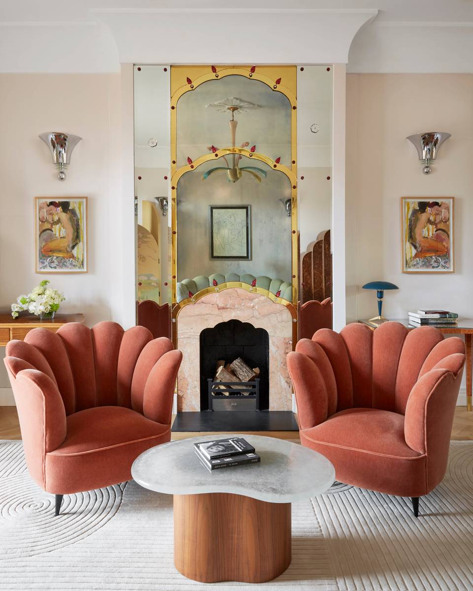 Two scalloped velvet armchairs and a sculptural coffee table face a decorative fireplace with mirrored Art Deco surround in an elegant, light-filled suite.