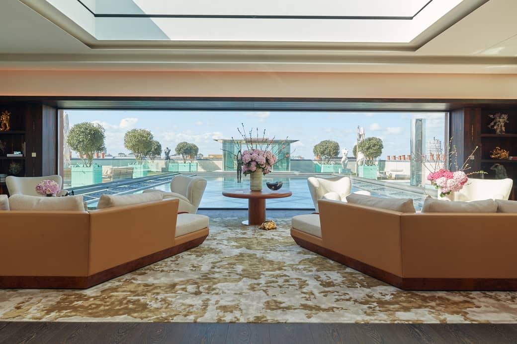 A luxurious sitting room features plush seating and elegant floral arrangements, offering a stunning view of a rooftop pool and manicured greenery under a bright sky.