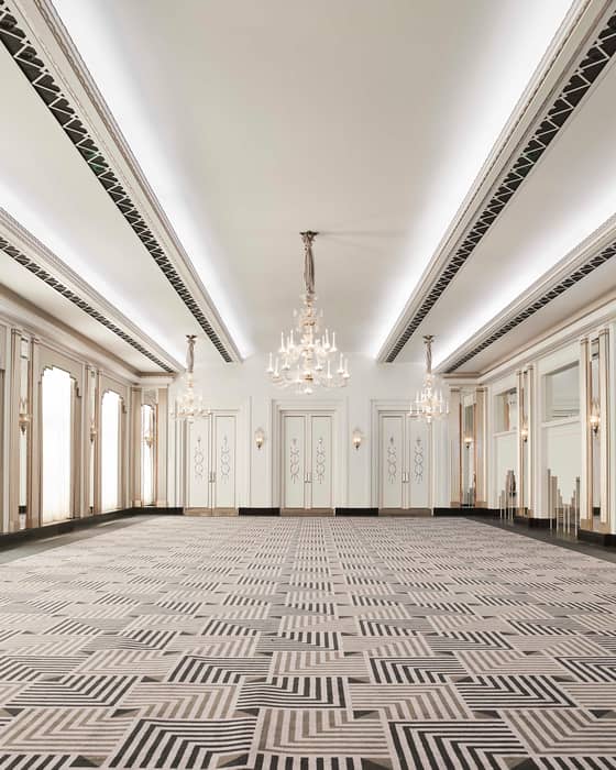 A majestic ballroom with crystal chandeliers, ornate mirrors, and geometric-patterned carpeting, bathed in natural light.