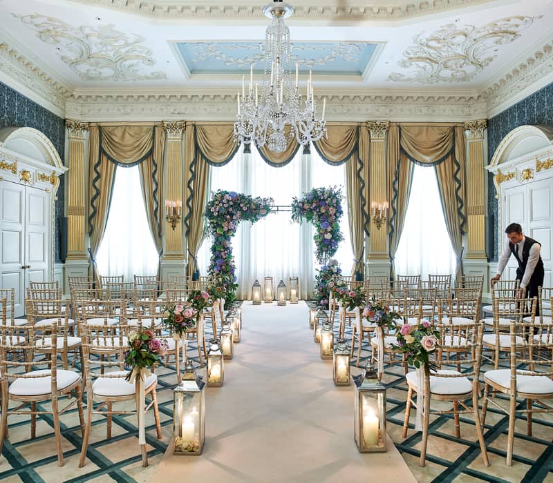 A grand and elegant ceremony setup in a luxurious drawing room with ornate decor, featuring a floral arch adorned with vibrant blooms, rows of gold chiavari chairs, and lanterns glowing with candlelight, as a staff member makes final adjustments.